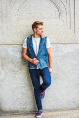 Young American Man with little beard, wearing blue Denim hoody sleeveless vest jacket, white T shirt, jeans, fashionable shoes, standing against vintage wall on street in New York, looking away. .