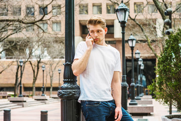 Young American College Student with little beard listening, talking on cell phone in New York, wearing white T shirt, standing against light pole on campus, looking away, waiting..