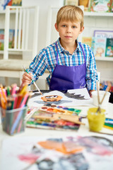 Wall Mural - Portrait of blonde little boy looking at camera while painting Halloween pictures sitting at table in art class