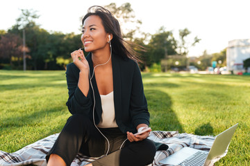 Poster - Young cheerful asian business woman listening to music, while resting after work in park