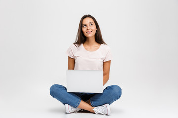 Wall Mural - Photo of young thinking brunette woman, holding and using laptop, while sitting with crossed legs