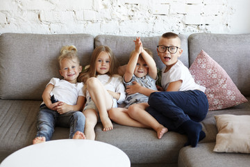 Group of four happy cute children boys and girls wearing casual clothes while having fun and relaxing together on couch, posing for common picture, looking at camera, smiling or making faces