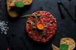 Beef tartar with a quail egg on top, grilled bread, greenery, served on a black stone plate.