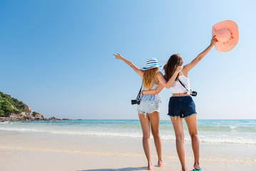 Portrait of two young asian female friends walking on the sea shore turn back at camera laughing. Multiracial young women strolling along a beach.