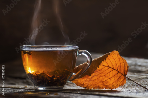 Zdjęcie XXL Przejrzysta filiżanka z herbatą i jesień żółtym suchym liściem na ciemnym tle. Dym kręci się z kubka. Jesienny nastrój przytulności i ciepła atmosfera.