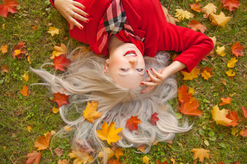 pretty smiling young blonde happy woman wearing red warm coat and lying on the green grass with yellow autumn leaves in the fall park alone top view