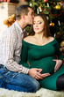 © Nestyda - Beautiful pregnant kissing couple in love on the background of a Christmas tree, having a rest on Christmas holidays before the new year. Big pregnant belly