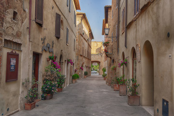 Wall Mural - Beautiful narrow street with sunlight and flowers in the small magical and old village of Pienza, Val D'Orcia Tuscany, Italy.