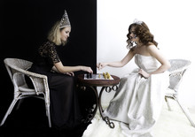 Two Girls Wearing Queen Dress; Diamond Crown And Other Jewelry Sitting In Chairs Opposite To Each Other And Playing Chess. 
Competition; Opposition Concepts. Black And White Background