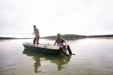 Two Men In Waders Are Fly Fishing From A Boat In A Lake.
