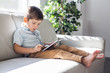 © Louis-Paul Photo - Little boy with digital tablet sitting on sofa, on home interior background