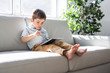 © Louis-Paul Photo - Little boy with digital tablet sitting on sofa, on home interior background
