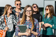 © proimagecontent - Cheerful brunette girl with white backpack holds books, glasses and headphones before modern building