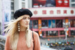 A beautiful young blonde woman dressed in a large warm pink sweater with a black beret and gorgeous earrings. She goes through a big spectacular Chinese city in autumn.
