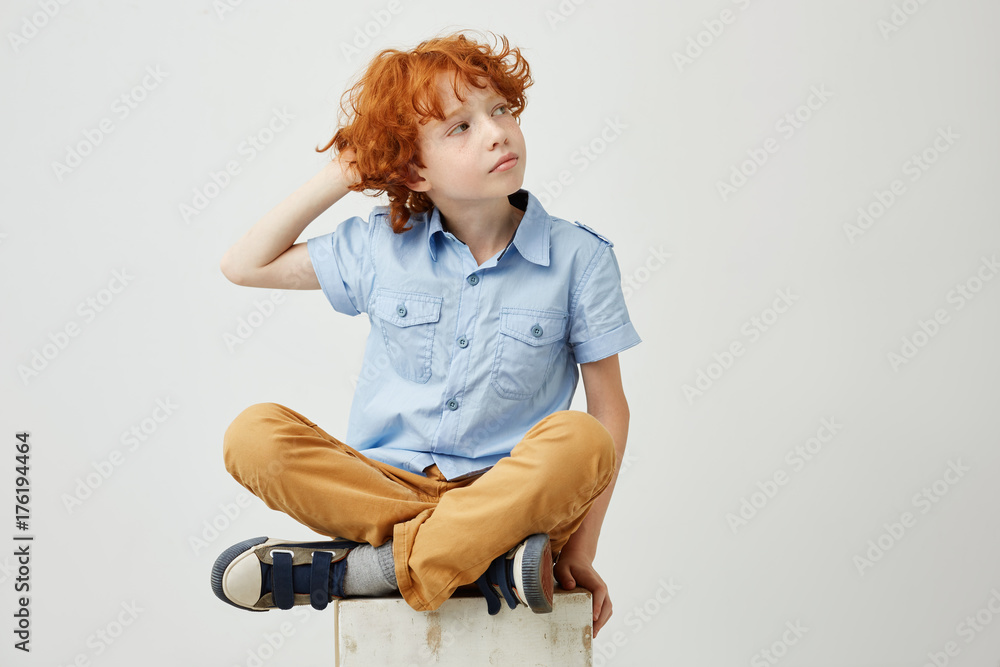 Beautiful ginger boy with curly hair and freckles in casual clothes holding hand on head, looking aside with relaxed expression.