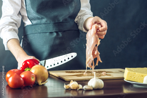 Zdjęcie XXL Szef kuchni w czarnym fartuszku wycina nożem filet z kurczaka. Koncepcja ekologicznych produktów do gotowania