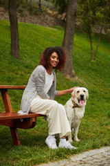 Wall Mural - Young african lady sitting on bench in park and holding dog