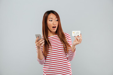 Wall Mural - Shocked asian woman in sweater using smartphone and credit card