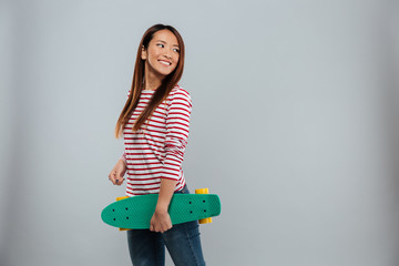 Poster - Side view of cheerful asian woman in sweater holding skateboard