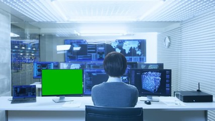 Fototapete - Back View of the IT Technician Working on with Neural Network on His Personal Computer with Green Screen Mock-up. He Works in a Big System Control Data Center with Multiple Monitors Showing Graphics.