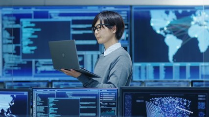 Wall Mural - In the System Control Room IT Technician Holds and Works on a Laptop, in Background Multiple Displays with Graphics.  Shot on RED EPIC-W 8K Helium Cinema Camera.