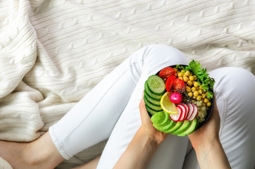 Girl in white jeans holds in hands fork, vegan breakfast meal in bowl with avocado, quinoa, cucumber, radish, salad, lemon, cherry tomatoes, chickpea, chia seeds. Top view, copy space. Clean eating.