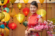 © DragonImages - Waist-up portrait of attractive young Asian woman wearing traditional costume Ao Dai posing for photography with sakura twig in hands, Chinese lanterns on background