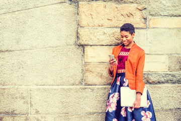 Unhappy Young African American Woman thinking outside in New York, wearing orange red jacket, flower patterned skirt, black boot shoes, holding laptop computer, standing by stone wall on campus, sad.