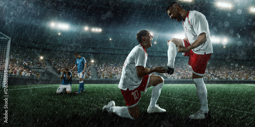 Plakat Piłkarze emocjonalnie szczęśliwy po zwycięstwie. Gracze radośnie krzyczą na dużym profesjonalnym deszczowym stadionie. Drużyna piłkarska nosi mundur niemarkowy.