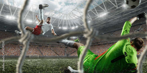 Plakat Piłkarz uderza piłkę do bramki na profesjonalnym stadionie. Bramkarz chroni bramę do piłki nożnej. Gracze mają na sobie niezarejestrowany mundur sportowy. Zobacz przez bramę piłki nożnej.
