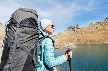 A hiker girl in sunglasses and a hat with a backpack and mountain gear with tracking treks in her hands looks at the beautiful view of a high mountain lake in an archipelago in the northern Caucasus