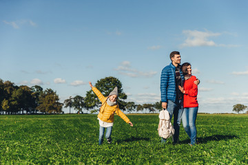 Playful little child runs on green meadow, plays near parents, who embrace each other, look into distance with thoughtful expressions. Family has walk or stroll in countryside, rest after noise city