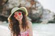 © Aleksandar Novoselski/Stocksy - Happy woman with hat and sunglasses at the beach, looking at camera
