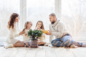 A happy family in white sweaters prepares to meet New Year in a beautiful loft with panoramic windows. Young parents with little daughters decorate a small Christmas tree in a pot. Copy space, bokeh