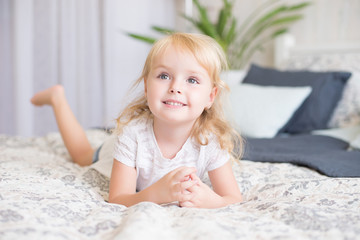 Cute pretty little girl with a beaming smile lying on her stomach a bed looking up into the air with a happy expression as she watches something.