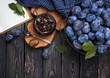 Homemade plum jam in a glass jar and fresh blue plums in a bowl on a dark rustic wooden background with copy space top view.