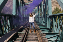 Young Beautiful Woman Standing On A Bridge And Railway. Holding A Purple Bomb Smoke. Sunny. Lifestyle