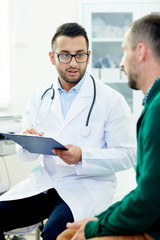 Wall Mural - Young clinician making notes in medical card of patient while talking to him