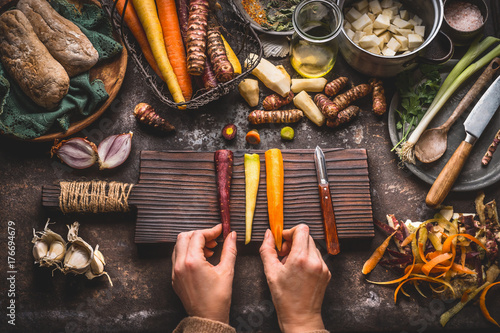 Zdjęcie XXL Zdrowe warzywa gotowania i jedzenie koncepcja. Żeńska kobieta wręcza trzymać kolorowe marchewki na kuchennego stołu tle z warzyw obieraczką, nożem, tnącą deską i kucharstwo garnkiem, odgórny widok