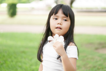 Wall Mural - Asian children cute or kid girl holding white plumeria flower and make face fragrance on nature garden and green grass with tree for holiday relax on soft focus