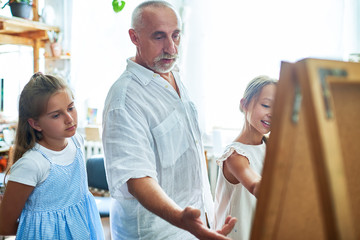 Wall Mural - Portrait of mature art teacher teaching children painting in art class explaining techniques and pointing at easel