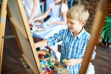 Wall Mural - Portrait of happy  little boy painting on easel during art class in sunlit studio enjoying work