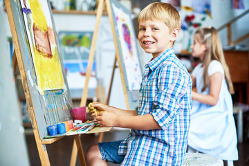 Wall Mural - Side view portrait of little boy fingerpainting house on easel in art studio and looking at camera smiling happily