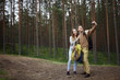 © Anatoliy Karlyuk - Cheerful young couple boy and girl tourists wearing hiking clothes and carrying backpacks smiling happily while taking self portrait on camera, standing on trail in forest with pines in background