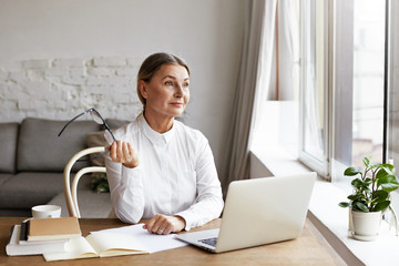 Beautiful mature female English teacher holding eyeglasses in hand and looking through window with thoughtful dreamy expression while sitting at desk in her study, doing paperwork and using laptop