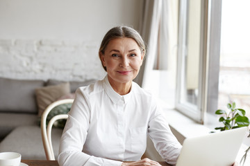 Wall Mural - Portrait of happy skilled middle aged woman life coach, business consultant, psychologist or medical advisor smiling joyfully at camera, working on laptop, enjoying her job, helping people online