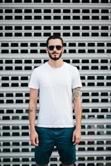 Wall Mural - A stylish man with a beard in a white T-shirt and glasses on a city background. Street photo