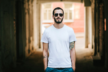 Wall Mural - Young stylish man wearing white blank t-shirt with beard in glasses, standing on the street on city background. Street photo