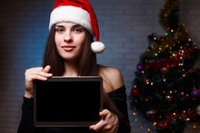 Young Beautiful Woman In Evening Dress And Santa Cap Holding A Tablet  With Empty Screen On Christmas Tree Background. New Year, Christmas, Gifts, Internet Shopping, Sales Concept