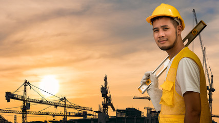 The portrait image of Asian young construction worker with leveling instrument in yellow helmet standing at the building construction site with background of tower crane operation during the sun set.